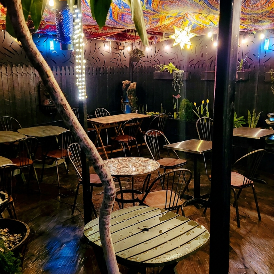 Eclectic outdoor restaurant patio with colorful mosaic canopy, string lights, plant-filled walls, fountain, and empty tables.