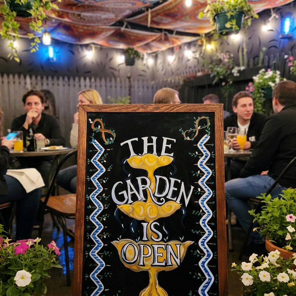 Outdoor restaurant garden patio with diners, hanging plants, colorful flowers, and a chalkboard sign reading “The Garden Is Open.”